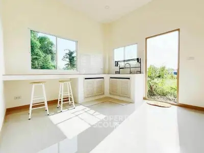 Bright kitchen with open layout and large windows overlooking greenery, featuring modern white cabinetry and bar stools.