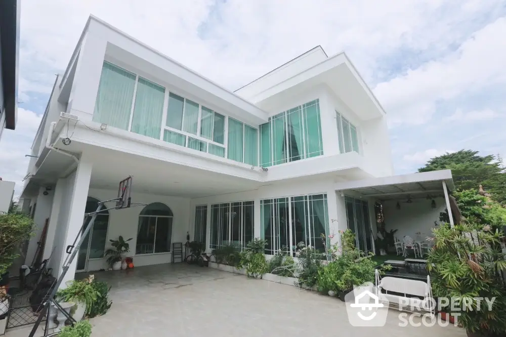 Modern two-story white house with large windows and a spacious patio, surrounded by lush greenery.