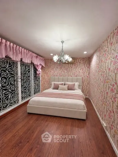 Charming bedroom with floral wallpaper and elegant chandelier, featuring a cozy bed and large windows with decorative grills.