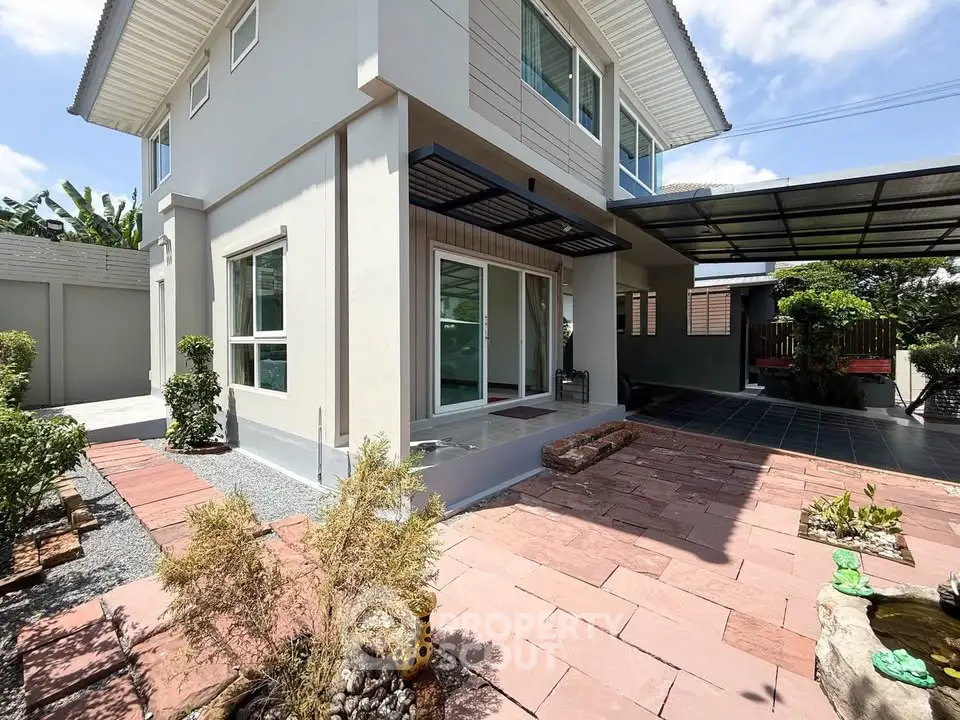 Modern two-story house with spacious patio and carport, surrounded by lush greenery and clear blue skies.