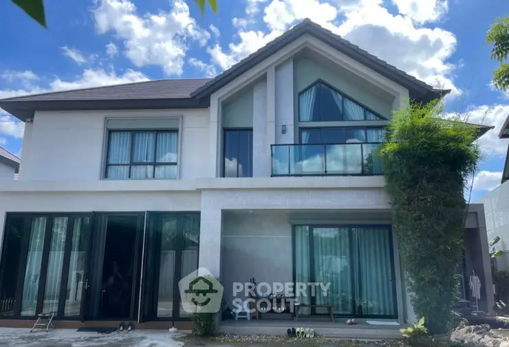 Modern two-story house with large windows and lush greenery under a bright blue sky.