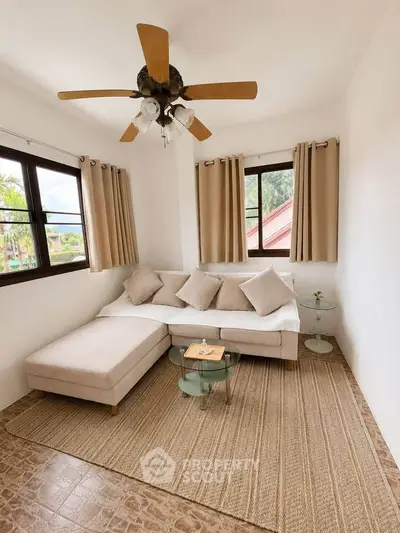 Cozy living room with beige sofa, ceiling fan, and natural light from large windows.
