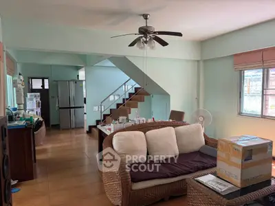 Spacious living room with wicker furniture and ceiling fan, featuring an open staircase and natural light.