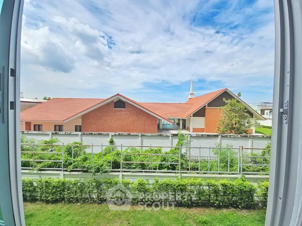 Stunning view from window overlooking lush garden and modern building with red roof under blue sky.