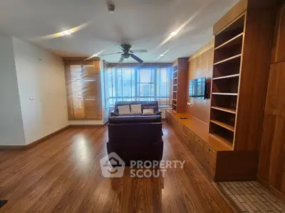Spacious living room with wooden flooring and built-in shelves, featuring a ceiling fan and large windows.
