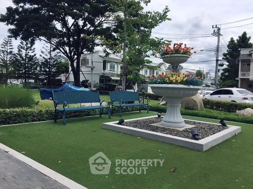 Charming community garden with vibrant green artificial turf, elegant benches, and a classic fountain centerpiece, surrounded by lush landscaping and residential backdrop.