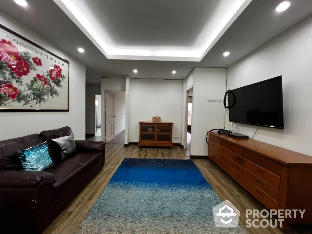 Spacious living room with modern lighting, plush seating, and a large flat-screen TV, complemented by a vibrant area rug and wooden accents.