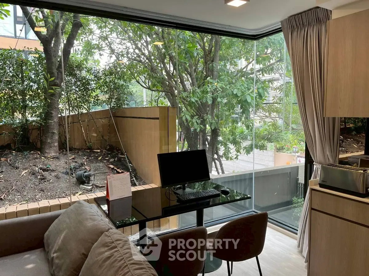 Modern living room with large glass windows and garden view, featuring a sleek workspace setup.