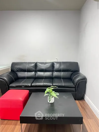 Modern living room with black leather sofa and red ottoman on wooden floor.