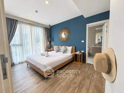 Modern bedroom with stylish decor and wooden flooring, featuring a sunburst mirror and large windows.