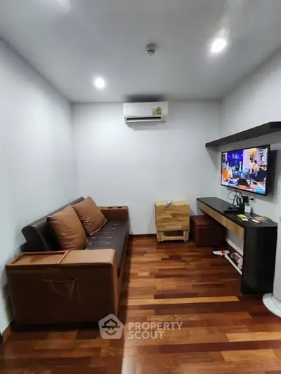 Cozy living room with modern furniture and wooden flooring, featuring a mounted TV and air conditioning.