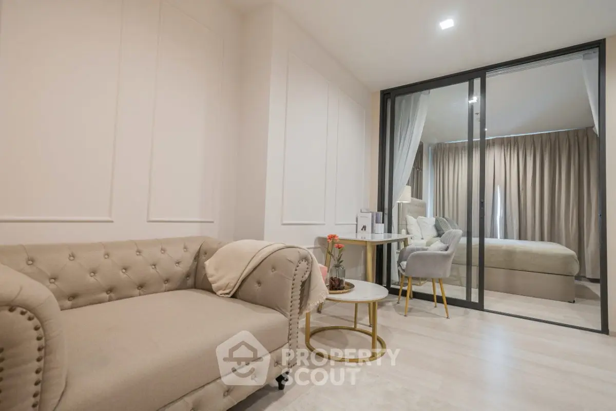 Elegant living room with beige sofa and glass partition leading to cozy bedroom.
