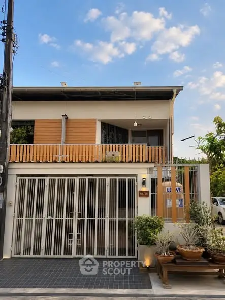 Modern two-story house with wooden accents and gated entrance, surrounded by lush greenery under a clear blue sky.