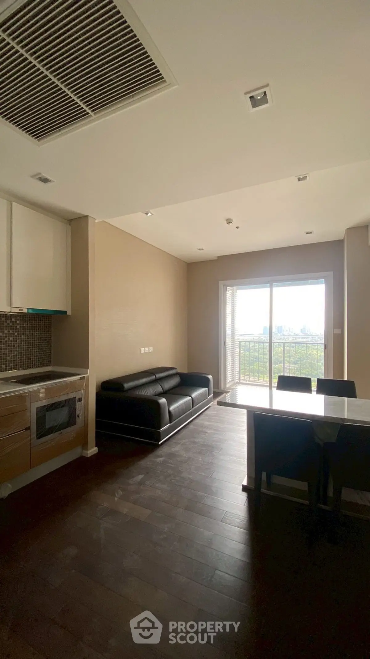 Modern living room with open kitchen and balcony view, featuring sleek black sofa and dining area.