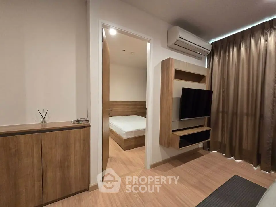 Modern living room with wooden furniture and a view into a cozy bedroom, featuring a mounted TV and air conditioning.