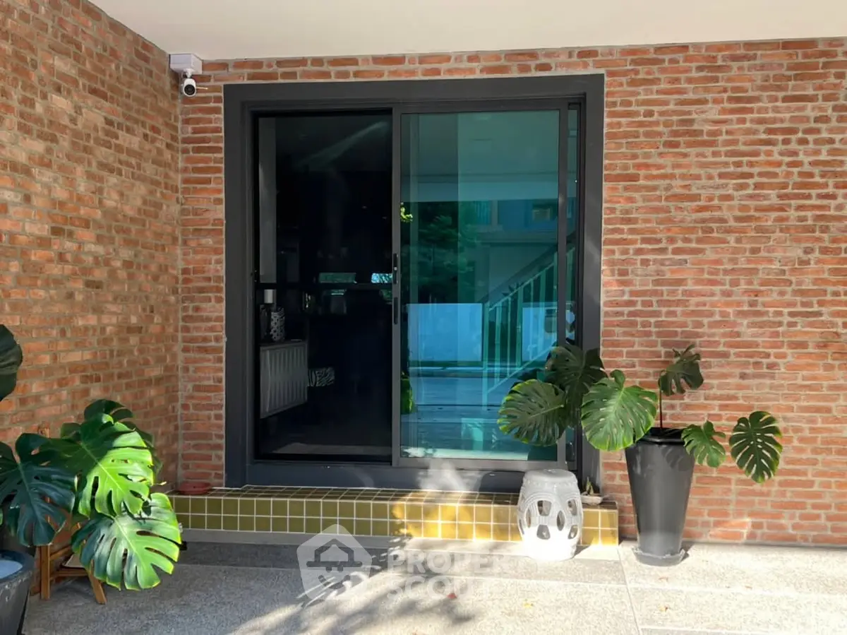 Modern entrance with brick wall and glass door, surrounded by lush potted plants.