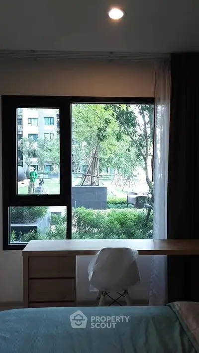 Cozy bedroom with a view of lush garden through large window, featuring a wooden desk and chair.