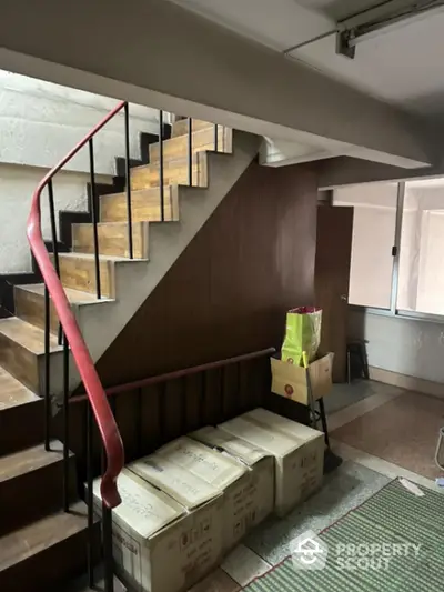 Spacious interior with a modern wooden staircase and a unique red handrail, complemented by natural light from the adjacent window.