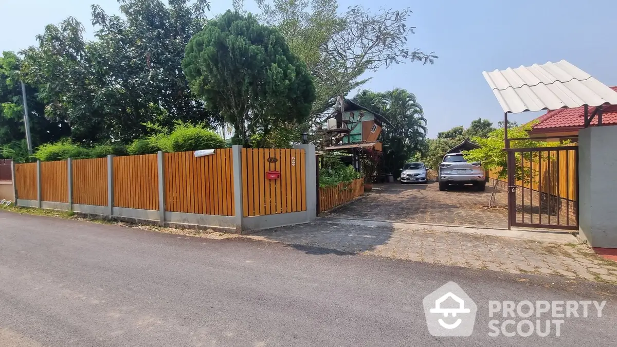 Charming property entrance with secure wooden gate, paved driveway, and lush greenery, offering a welcoming approach to a serene suburban home.