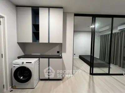 Modern apartment interior with washing machine and sliding glass doors leading to a bedroom.