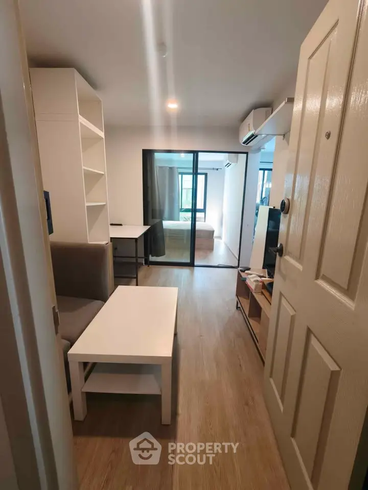 Modern apartment entrance with open living area and view into bedroom, featuring sleek furniture and wooden flooring.