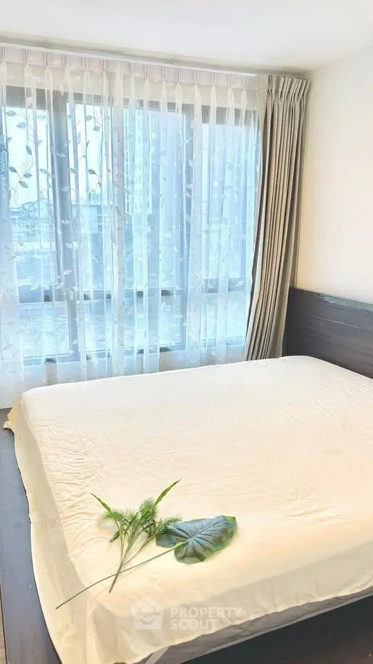 Bright bedroom with large window and elegant curtains, featuring a cozy bed and decorative plants.