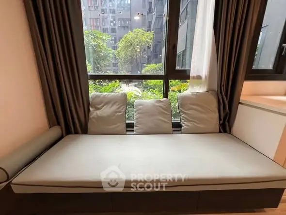 Cozy window seat with cushions overlooking lush greenery in urban setting.