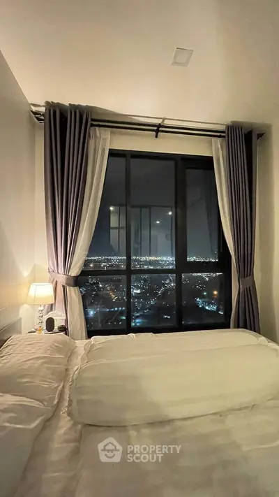 Cozy bedroom with stunning city night view through large window, perfect for urban living.
