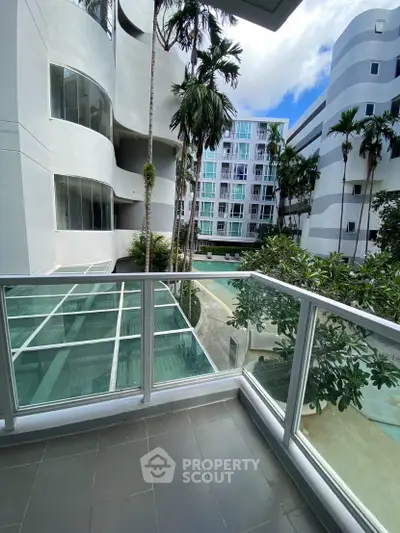 Modern apartment balcony with glass railing overlooking pool and lush palm trees.