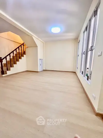 Spacious living room with wooden flooring and staircase, ample natural light from large windows.