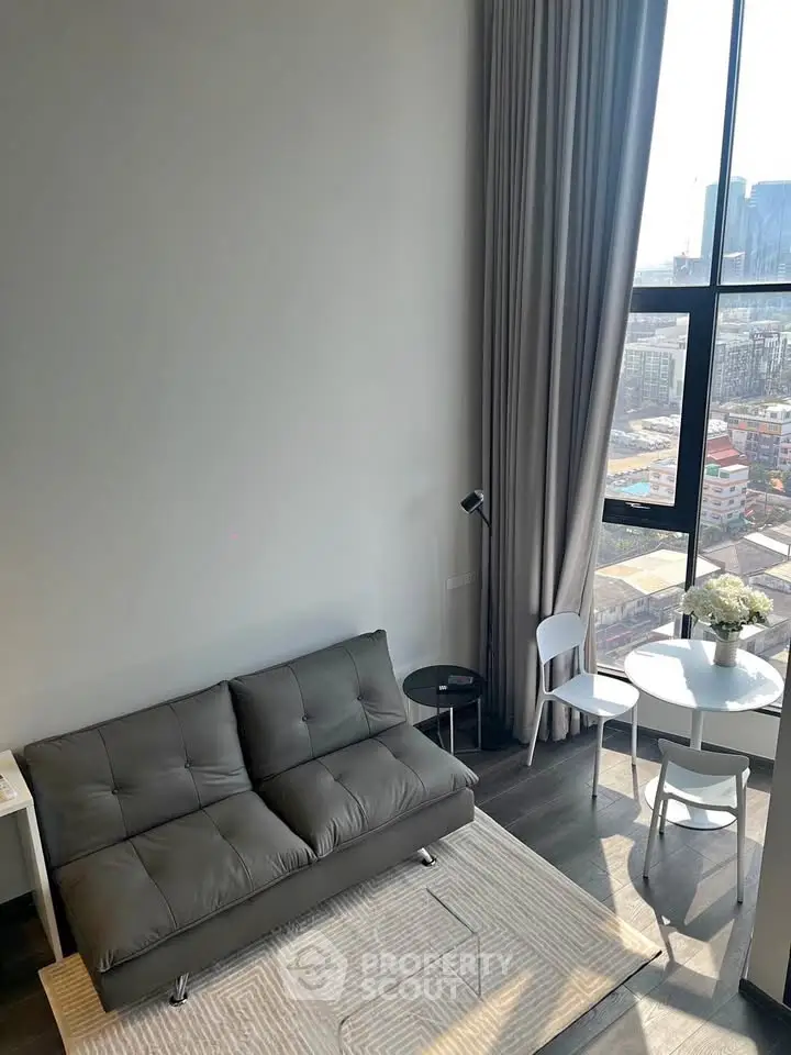 Modern living room with high ceiling and large windows offering city views, featuring a sleek sofa and dining area.