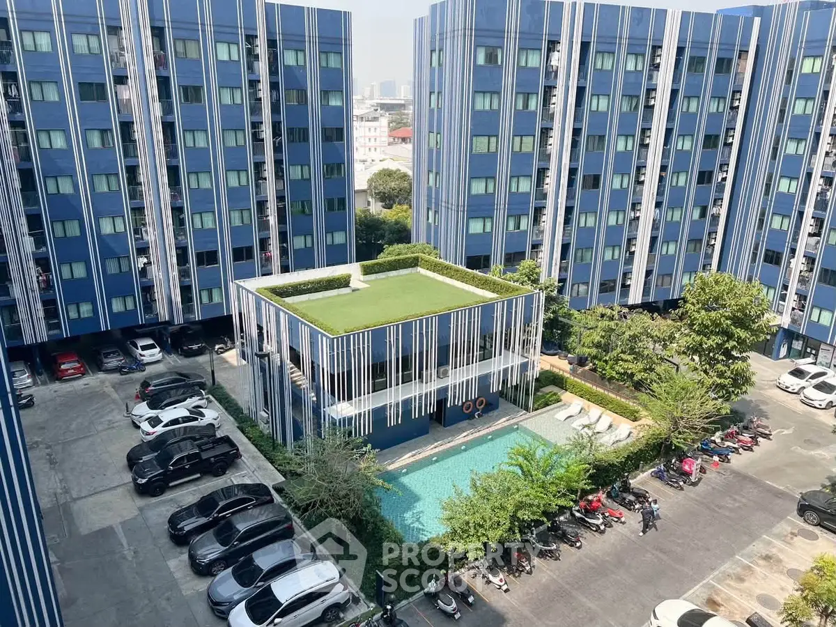 Modern apartment complex with pool and parking area, featuring blue facade and lush greenery.