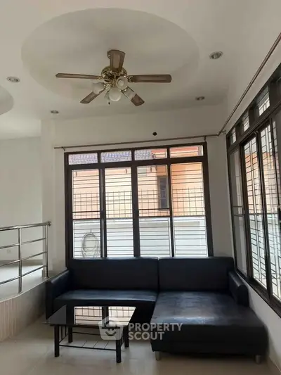 Spacious living room with modern black sofa and ceiling fan, large windows for natural light.