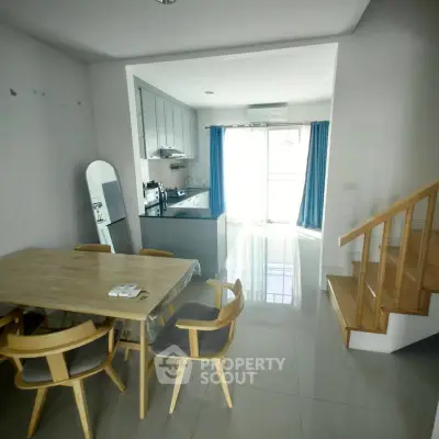 Modern open-plan living area with dining table and kitchen, featuring natural light and wooden staircase.