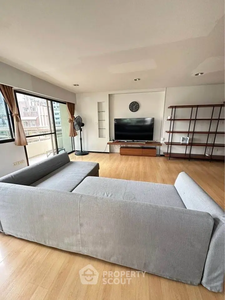 Spacious living room with modern gray sofa and large TV, featuring wooden flooring and ample natural light.