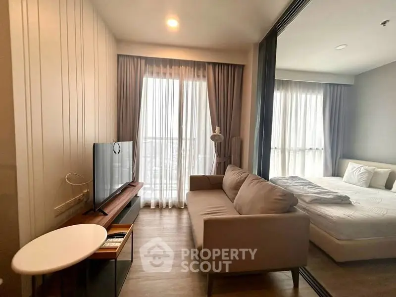 Modern living room with cozy sofa and TV, adjacent to a bright bedroom with large windows.