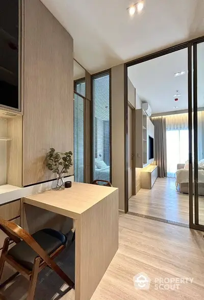 Modern study area with wooden desk and chair, leading to a spacious living room with natural light.