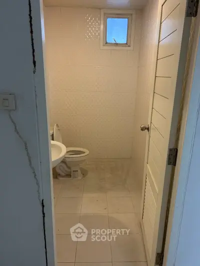Compact bathroom with white tiles and small window, featuring a toilet and sink.