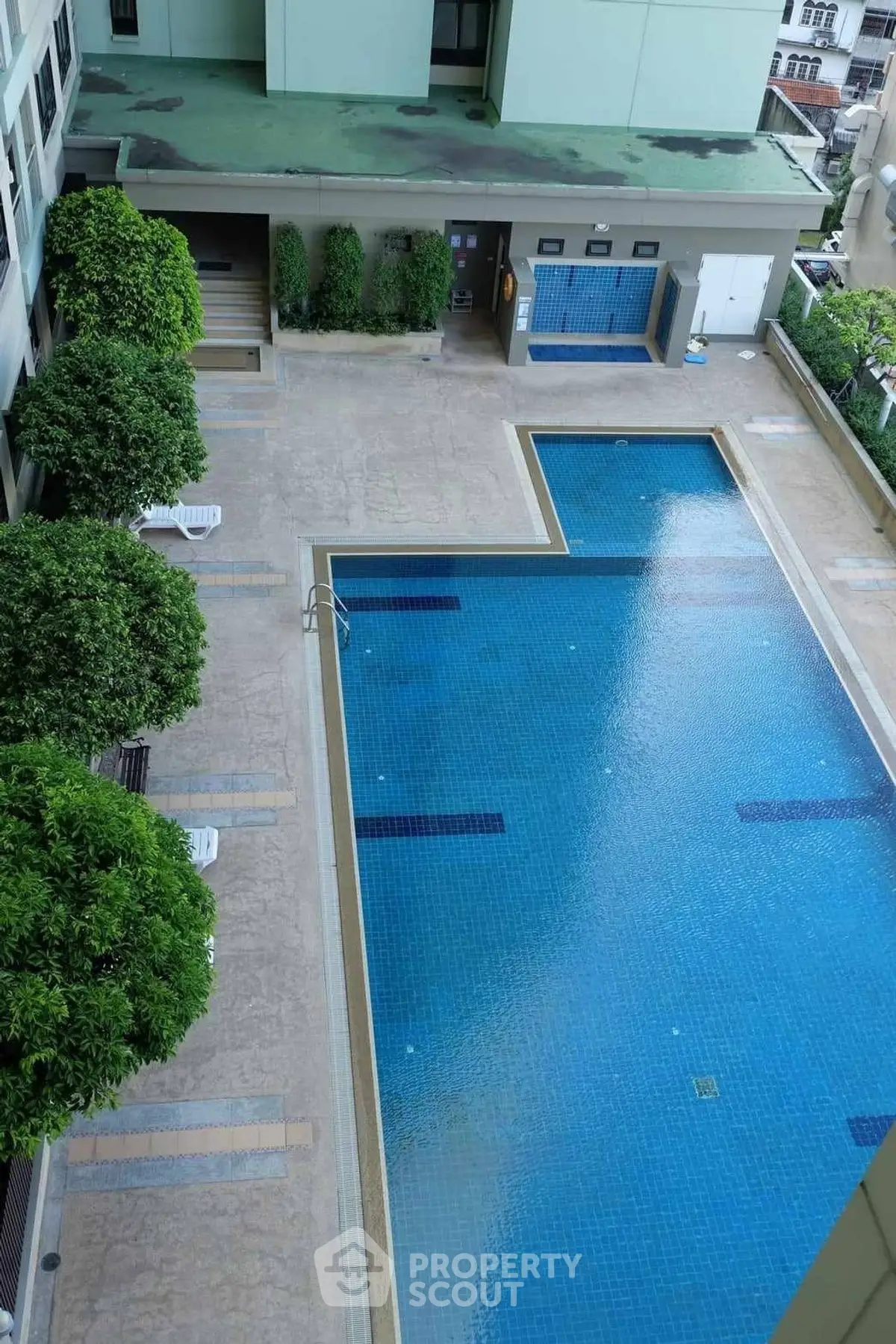 Spacious outdoor swimming pool in modern residential complex with lush greenery and seating area.