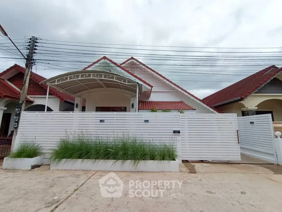 Charming single-story house with red roof and white fence, featuring a covered entrance and lush greenery.