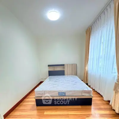 Cozy bedroom with wooden flooring and large window with curtains, featuring a comfortable bed.