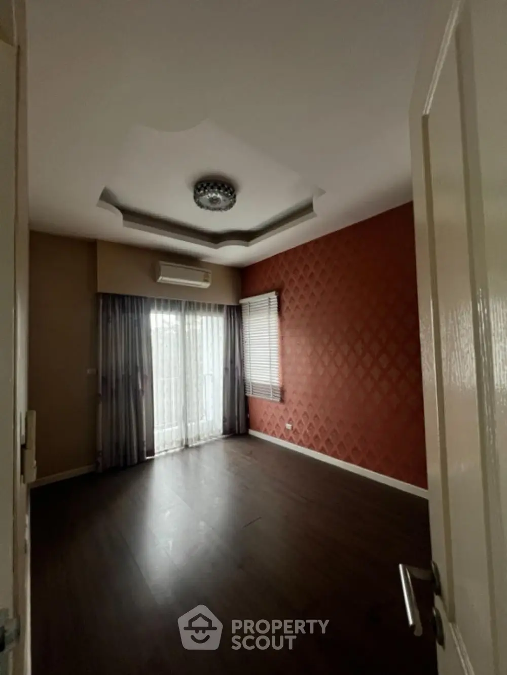 Spacious living room with elegant dark flooring, textured red accent wall, and sophisticated ceiling design, complemented by ample natural light.