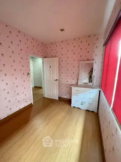 Charming bedroom with floral wallpaper and natural light, featuring a dresser and wooden flooring.