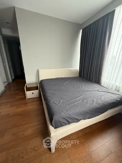Spacious bedroom with elegant wooden flooring and large window draped with curtains.