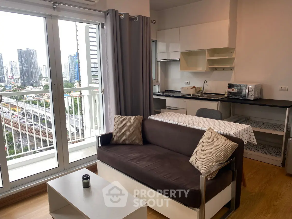 Modern apartment living room with balcony and city view, featuring a cozy sofa and compact kitchen area.