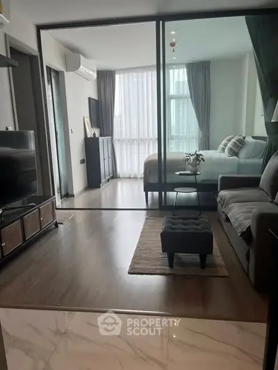 Modern living room with glass partition leading to a cozy bedroom, featuring stylish decor and natural light.