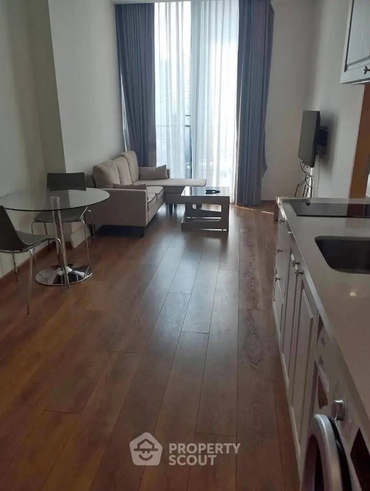Modern living room with open kitchen, featuring hardwood floors and natural light from large windows.