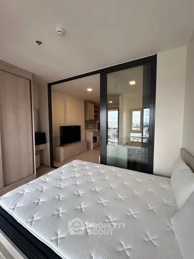Modern bedroom with sliding glass doors leading to a stylish living area, featuring a sleek TV setup and kitchen view.