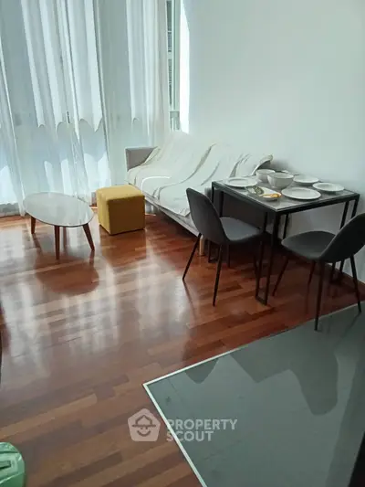 Modern living room with wooden flooring, cozy sofa, dining table, and natural light through large windows.