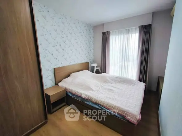 Cozy bedroom with wooden furniture and floral wallpaper, featuring a large window with curtains.
